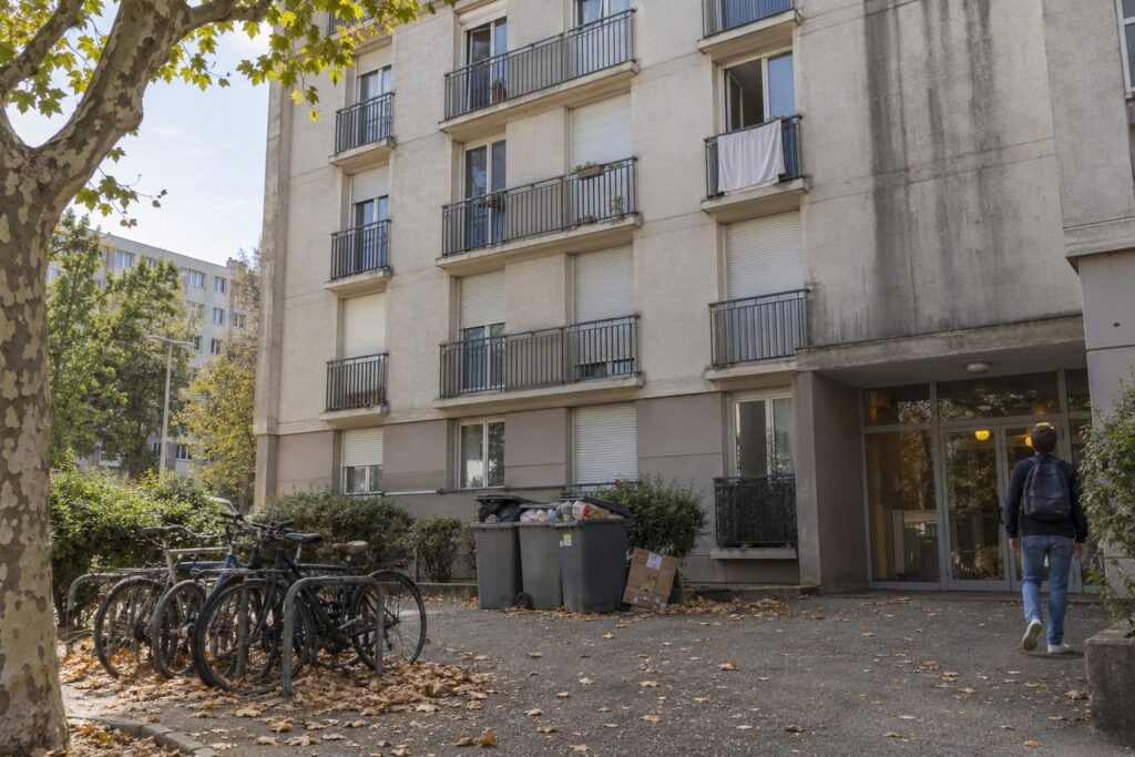 Façade d'une résidence étudiante des années 1970 avec vélos, poubelles et un étudiant de dos près de l'entrée