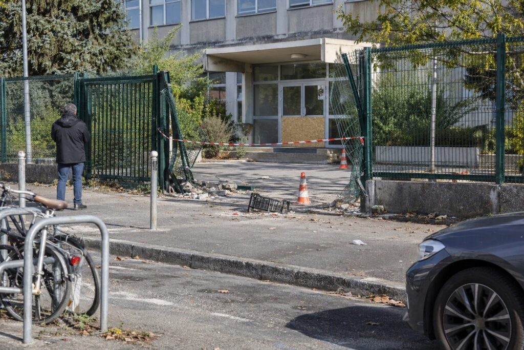 Section de grille de lycée tordue et rubalisée sur un trottoir ensoleillé, un adulte au loin de dos