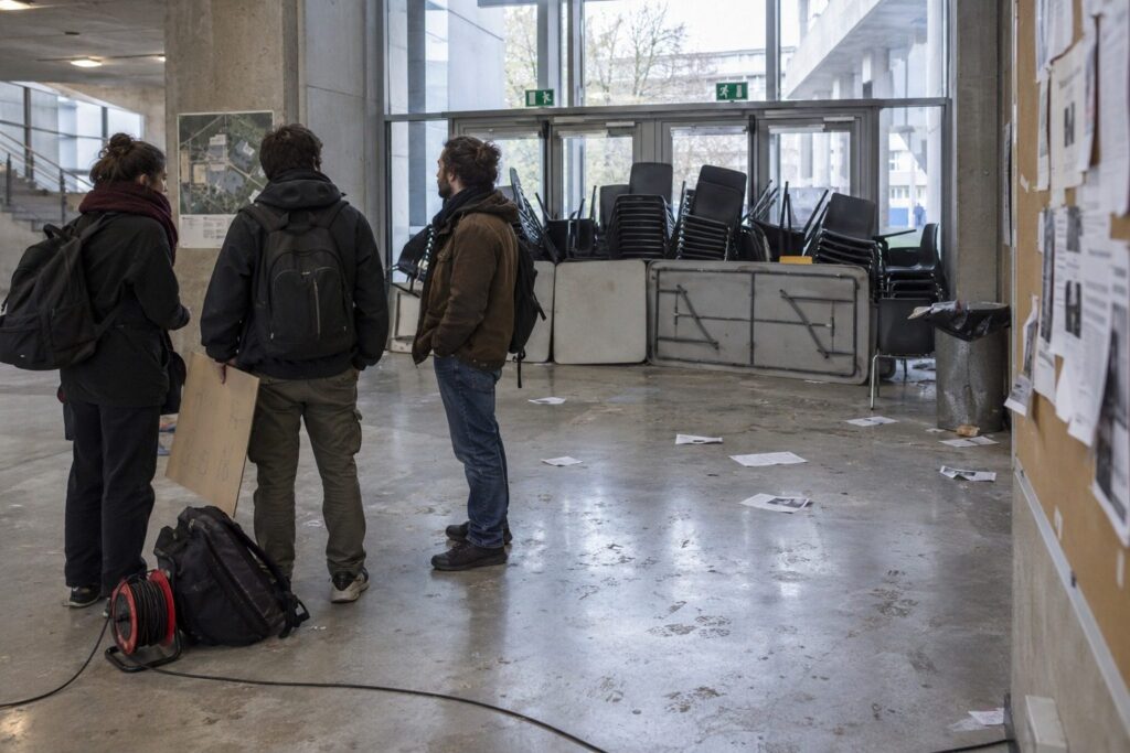 Entrée d'un bâtiment universitaire bloquée par des chaises empilées, quelques étudiants adultes de dos devant les portes vitrées